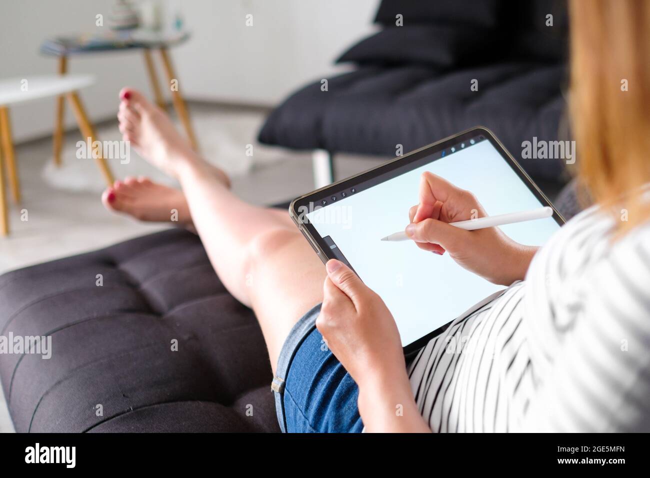 Frau, die mit Bleistift auf dem Tablett arbeitet oder zeichnet und auf dem Sofa liegt. Ein Designer oder Künstler arbeitet von zu Hause aus Stockfoto