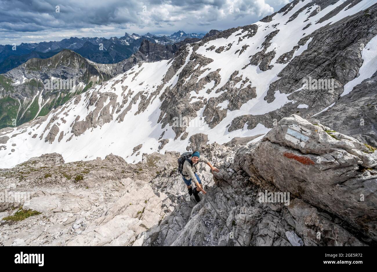 Wanderer bergab felsiges Gelände, Wanderweg mit einem Stahlseil gesichert, Bergpanorama mit alten Schneefeldern und felsigen Berggipfeln in der Stockfoto