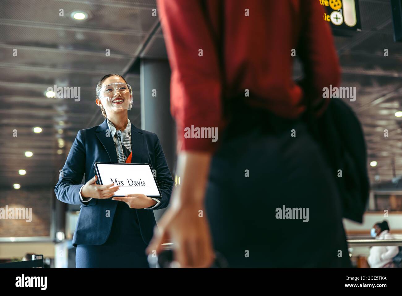 Der Fahrer begrüßt die Geschäftsfrau mit einem Schild am Ankunftsterminal. Chauffeur trägt Gesichtsschutz und hält die Beifahrer-Informationafel. Stockfoto