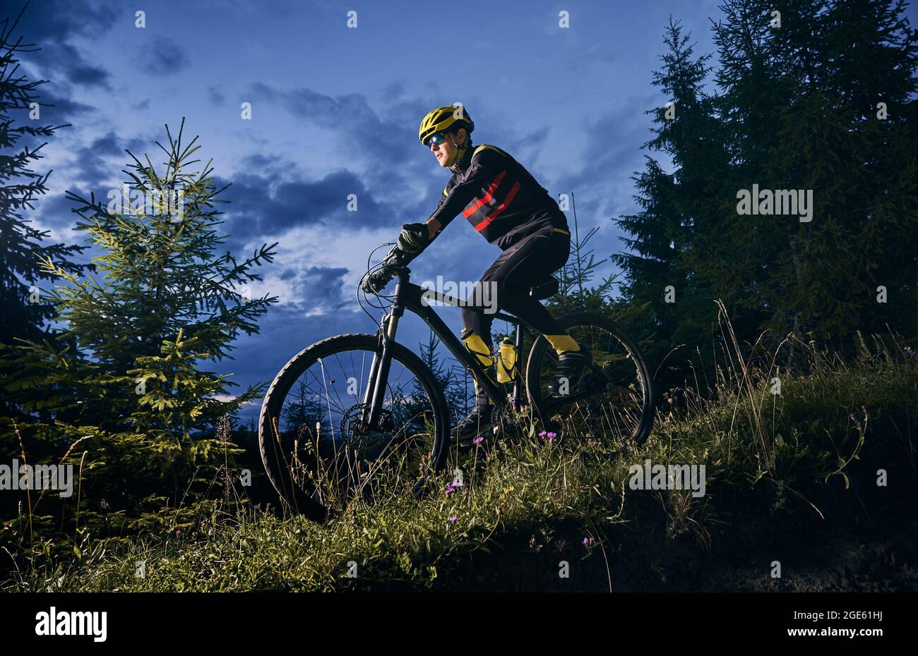 Junger Mann fährt Fahrrad bergab mit schönen blauen Abendhimmel auf dem Hintergrund. Männlicher Radfahrer im Sportfahranzug, der nachts den grasbewachsenen Hügel hinunterradelt. Konzept von Sport, Radfahren und aktiver Freizeit. Stockfoto