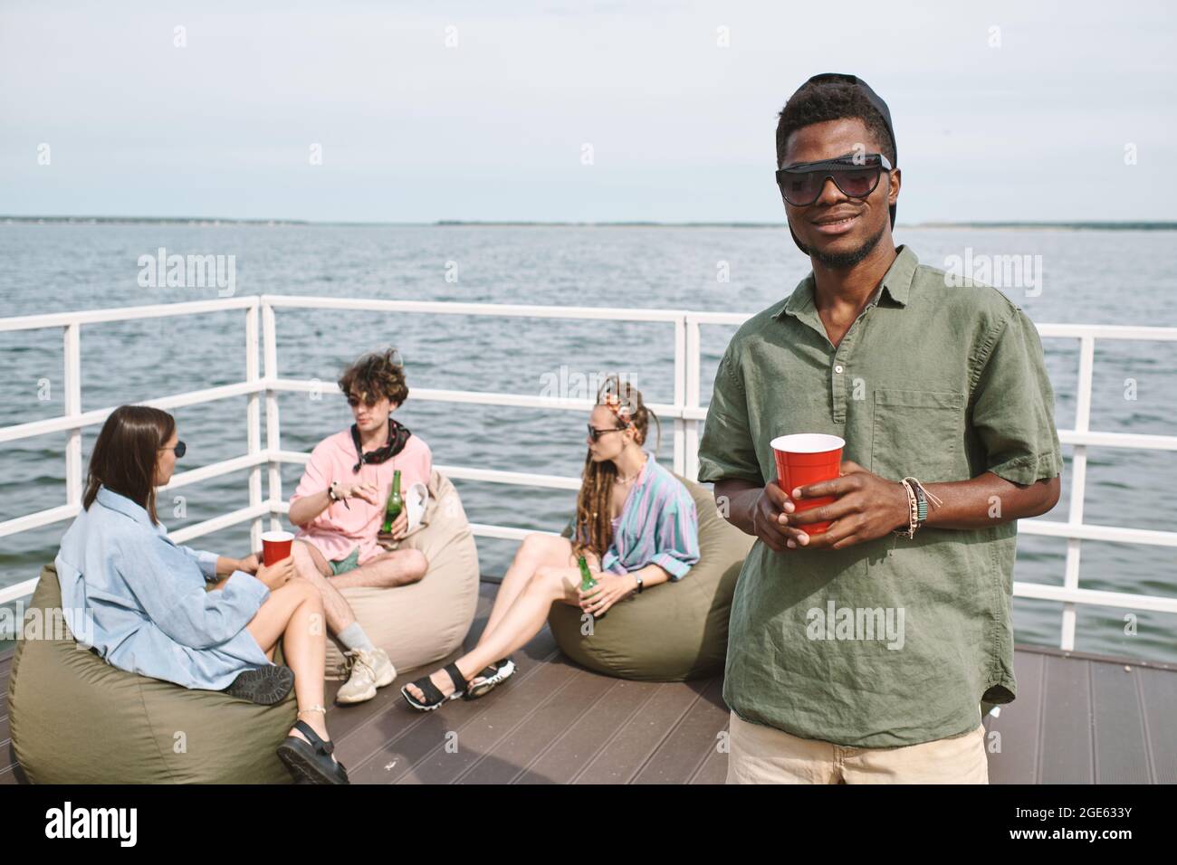 Junger schwarzer Mann, der auf einer Party am hölzernen Pier am See teilnimmt, trinkt Alkohol und schaut auf die Kamera Stockfoto