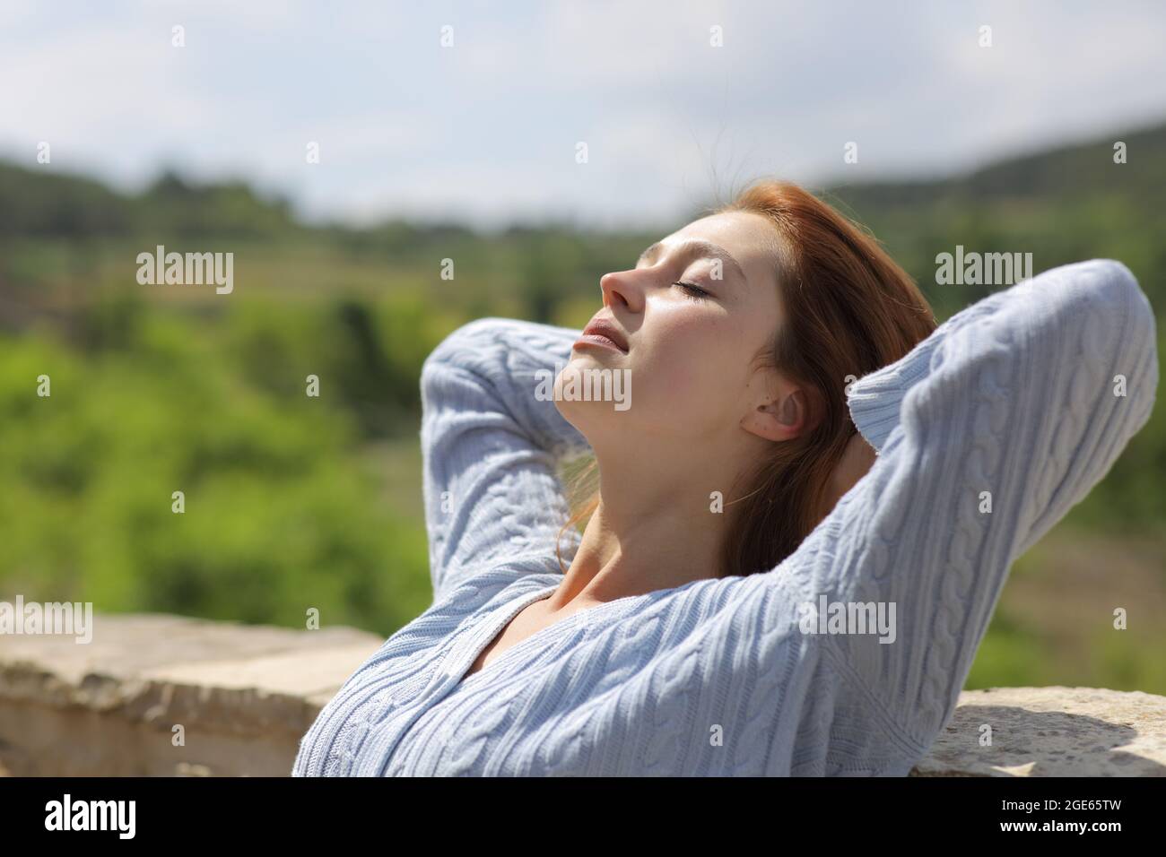 Entspannte Frau, die mit den Armen auf dem Kopf auf einem Balkon eines Landhauses sitzt Stockfoto