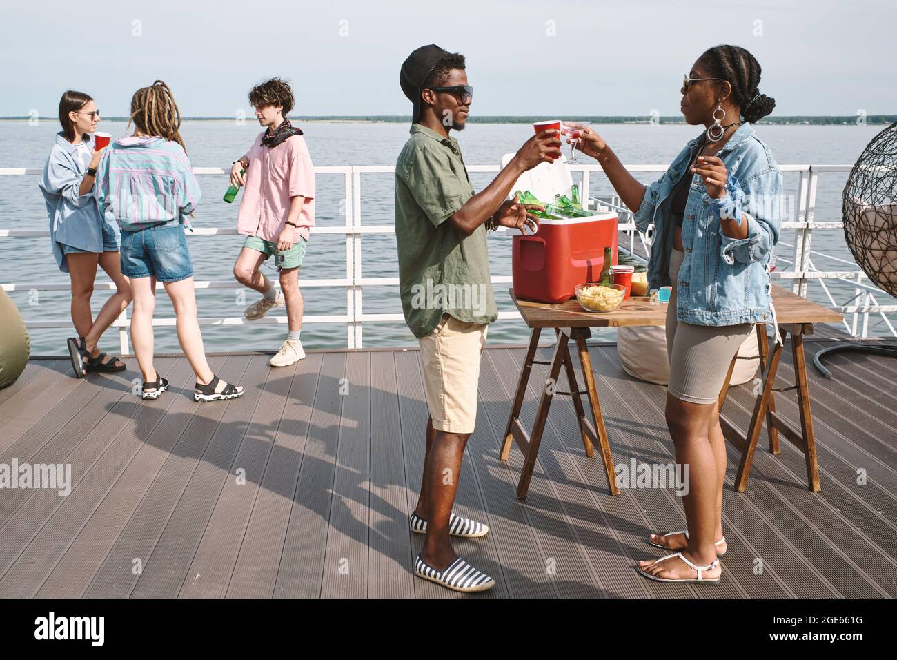 Junge Leute toasten mit erfrischenden Getränken, wenn ihre Freunde im Hintergrund auf der Party am Pier reden Stockfoto