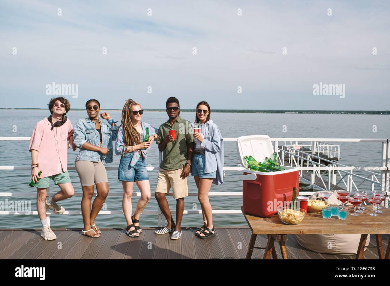 Fröhliche Freunde, die eine kleine Party auf dem hölzernen Pier mit Getränken und Snacks arrangieren Stockfoto