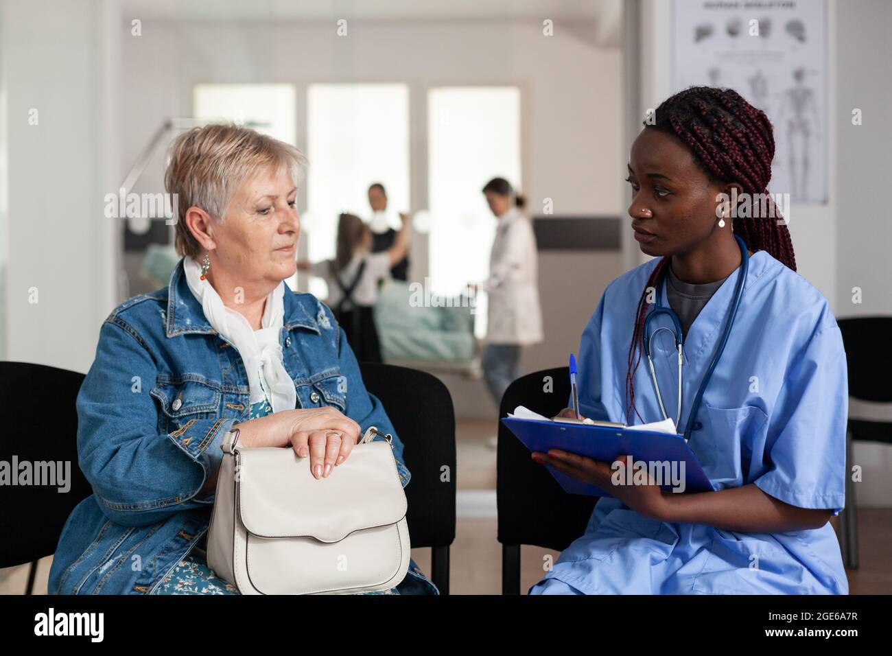 afroamerikanische Assistentin, die Krankheitssymptome mit älteren kranken älteren Patienten diskutiert und medizinische Behandlung im Wartezimmer des Krankenhauses auf die Zwischenablage schreibt. Support-Service im Gesundheitswesen Stockfoto