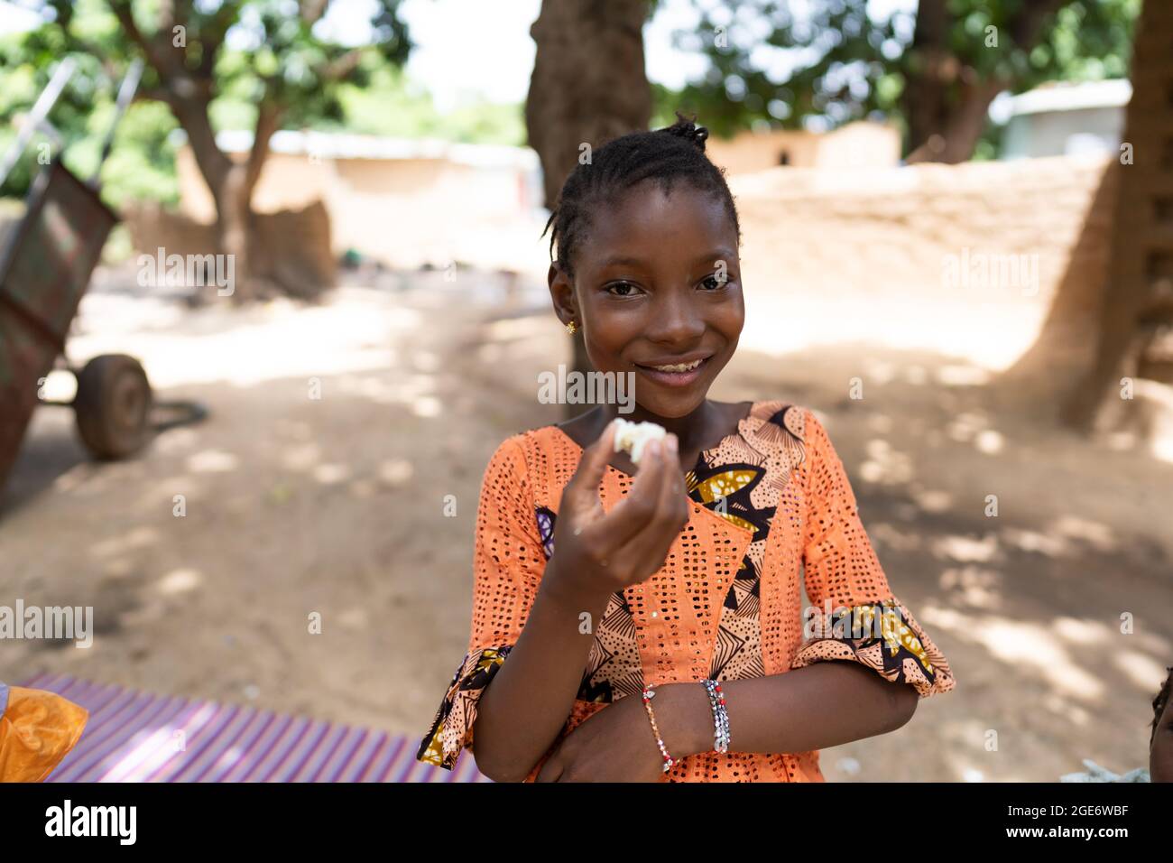 Stilvolles, schwarz-afrikanisches Mädchen, das im Hof ihrer Familie steht und vor der Kamera grinst, während es ein gekochtes Ei isst Stockfoto