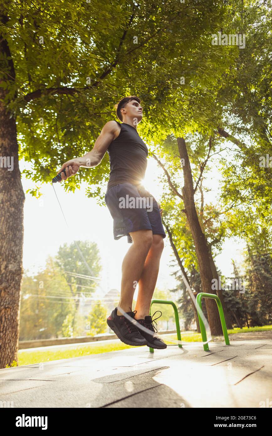 Ein junger, hübscher Kaukasusmann in Sportkleidung, der am hellen Sommertag mit einem Seil auf dem Sportplatz springt. Stockfoto