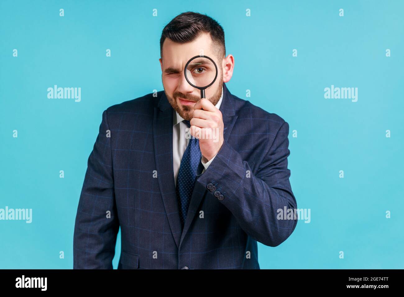 Junger, hübscher Geschäftsmann im dunklen offiziellen Stil, der eine Lupe hält und mit einem großen Zoomauge die Kamera anschaut. Innenaufnahme im Studio Stockfoto
