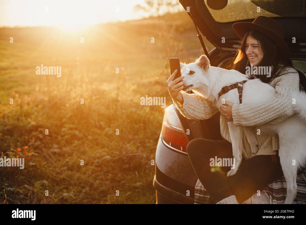 Stilvolle Hipster Frau, die Selfie-Foto mit niedlichen Hund im Kofferraum in warmen Sonnenuntergang Licht. Herbstlicher Roadtrip mit Haustier. Junge Frau mit Telefon und tra Stockfoto