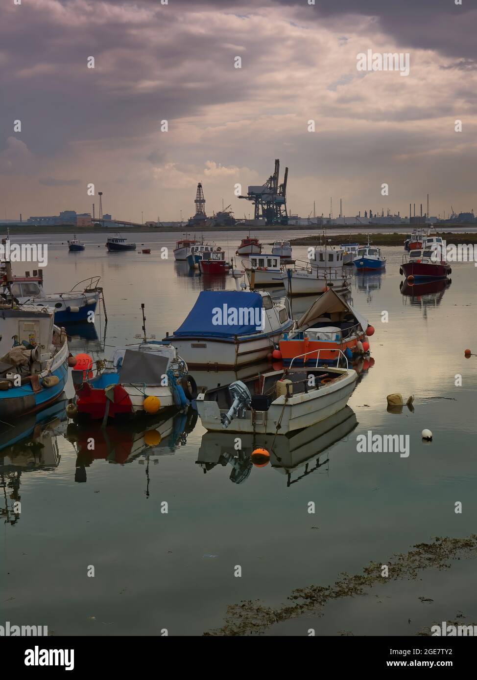 Die kleinen Fischerboote ruhen in den glasigen Gewässern vor dem industriellen Horizont, unter dem bedrohlichen Himmel. Stockfoto