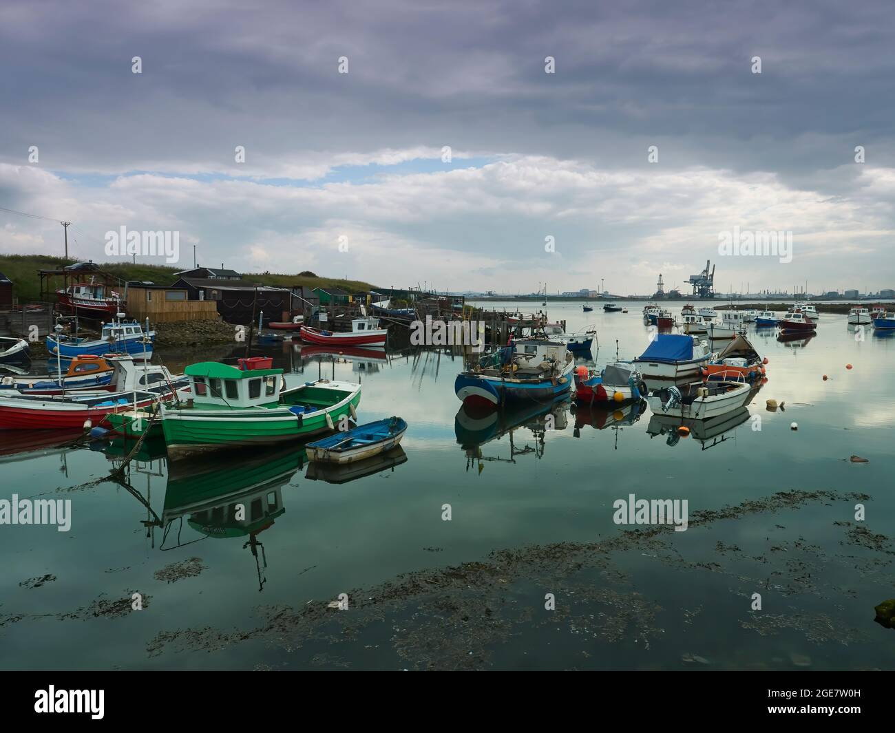 Fischerboote ruhen in den glasigen Gewässern von Paddy’s Hole, im Hintergrund die riesigen Containerkrane von Teesport. Stockfoto