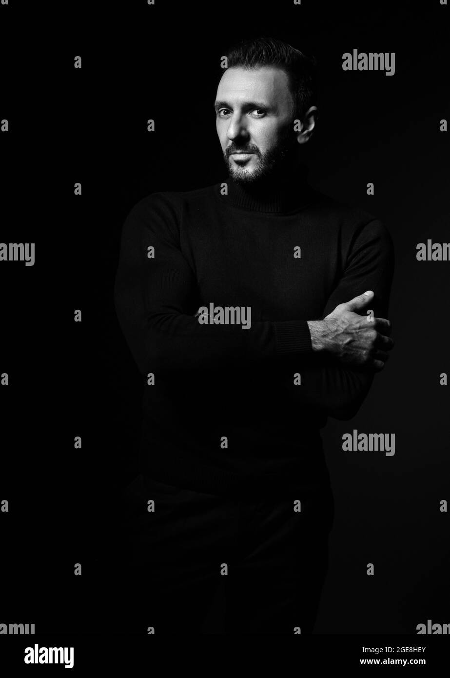 Schwarz und Weiß. Erwachsener bärtiger Mann im schwarzen Rollkragenpullover steht mit gekreuzten Armen auf der Brust und schaut auf die Kamera Stockfoto