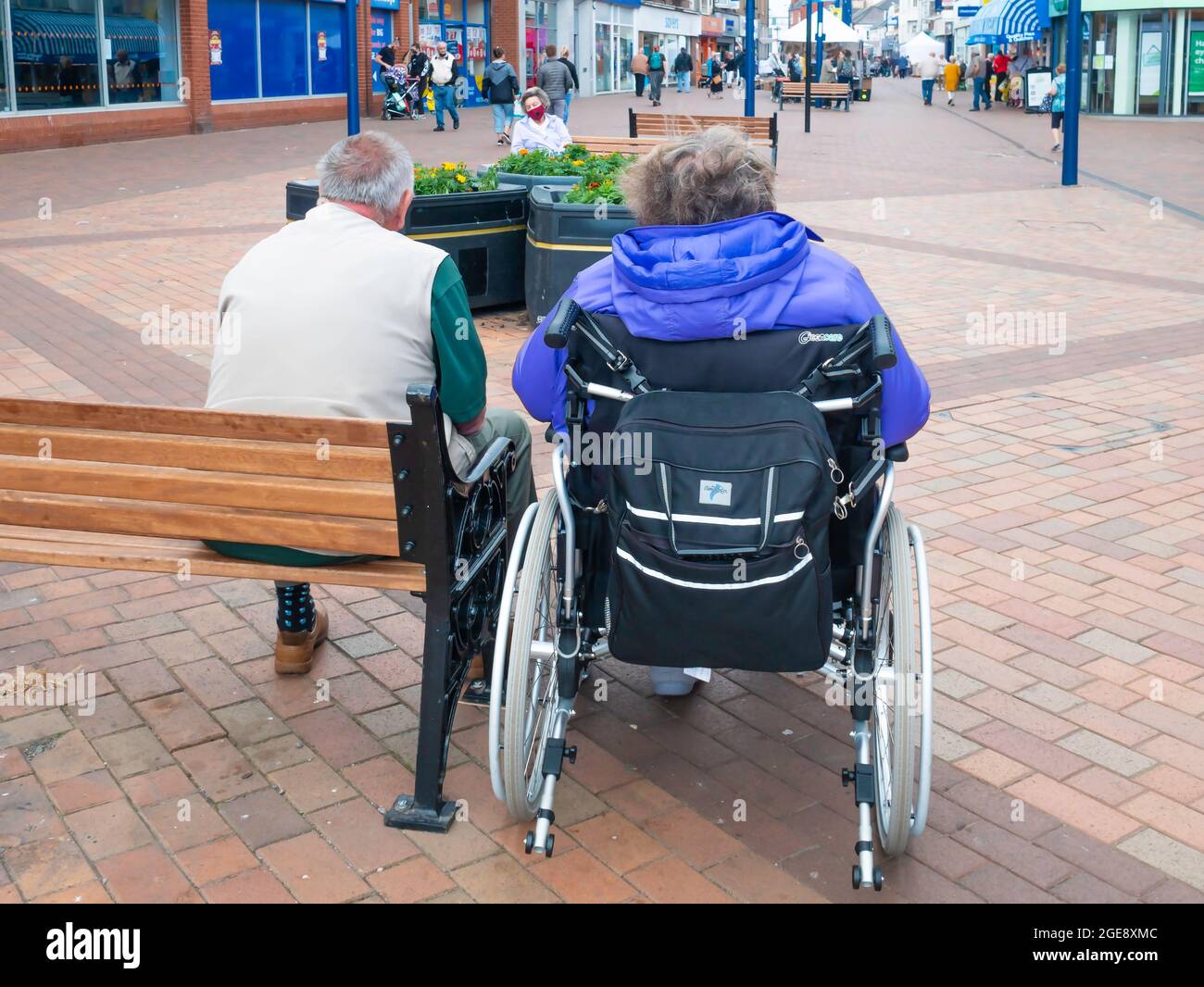 Das ältere Paar sitzt auf einer Bank, und sie genießt in einem eleganten Rollstuhl in Redcar High Street Cleveland, Großbritannien, ruhig die Sonne Stockfoto