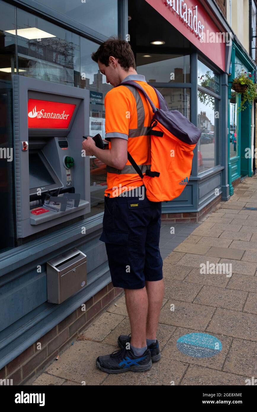 Ein großer junger Mann mit orangefarbenem Hemd und Rucksack, der an einem Geldautomaten von Santander teilhat Stockfoto