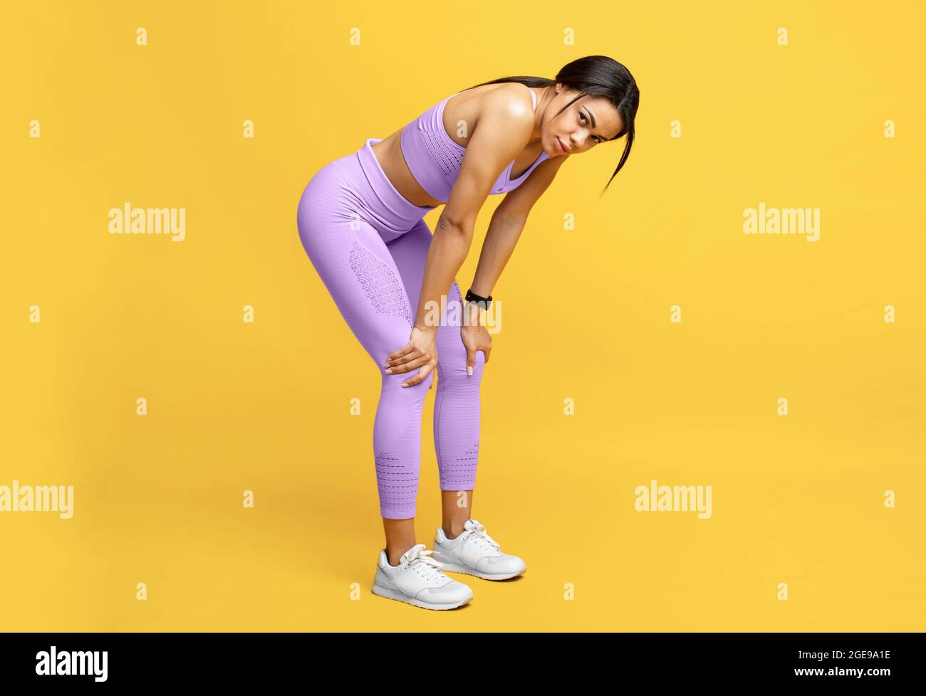 Müde afroamerikanische Dame, die beim Training Pause macht, sich ausruht und sich auf den Knien über dem gelben Studiohintergrund lehnt Stockfoto