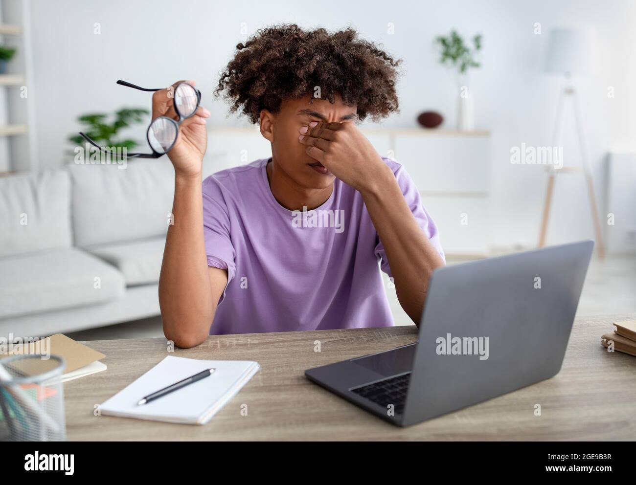 Schwarzer Teenager, der müde Augen reibt, mit einem Laptop am Schreibtisch sitzt, erschöpft vom Online-Studium Stockfoto