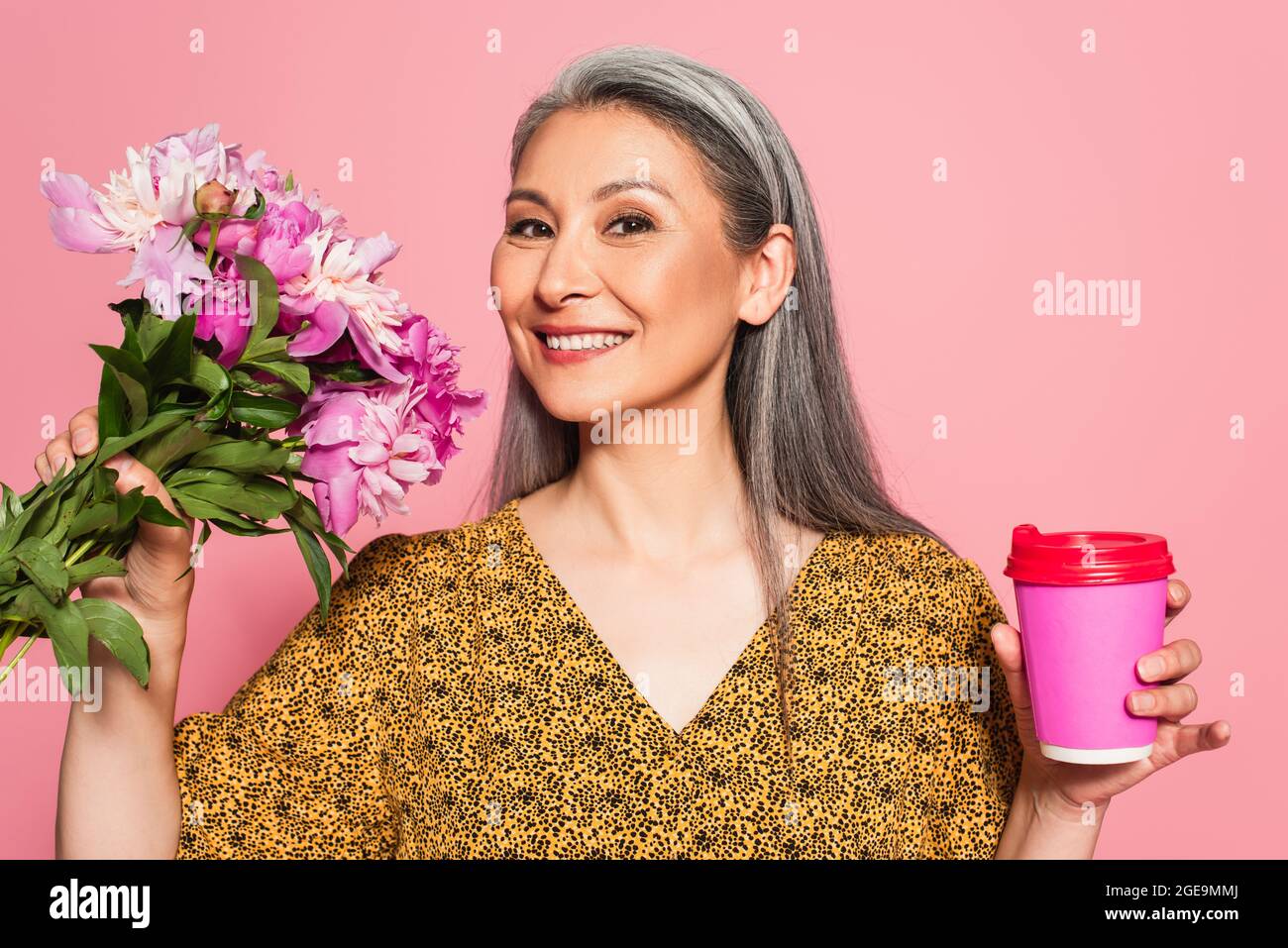 Fröhliche Frau mit Blumen und Kaffee zu gehen Blick auf Kamera isoliert auf rosa Stockfoto