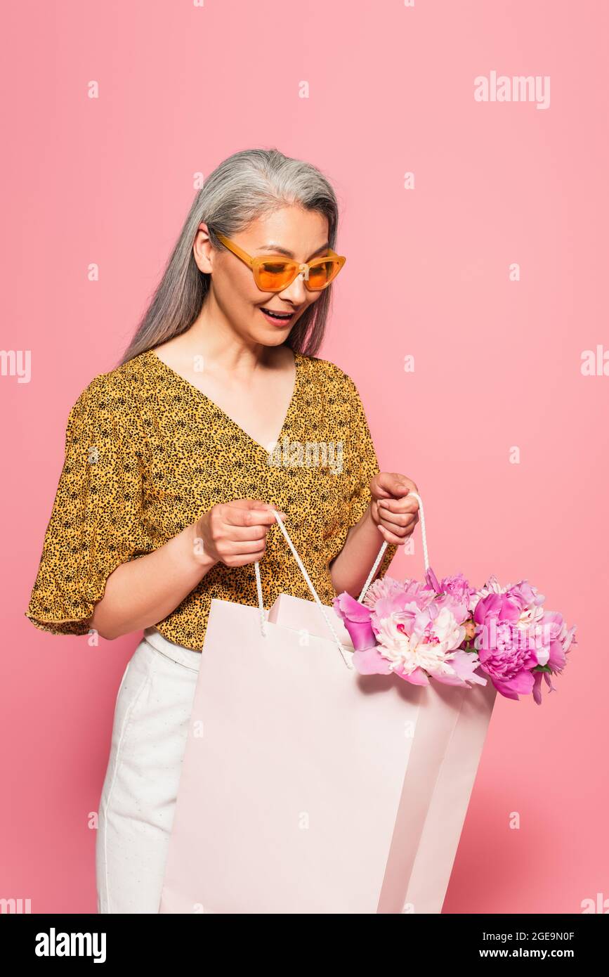 Erstaunt reife Frau Blick in Einkaufstasche mit Pfingstrosen isoliert auf rosa Stockfoto