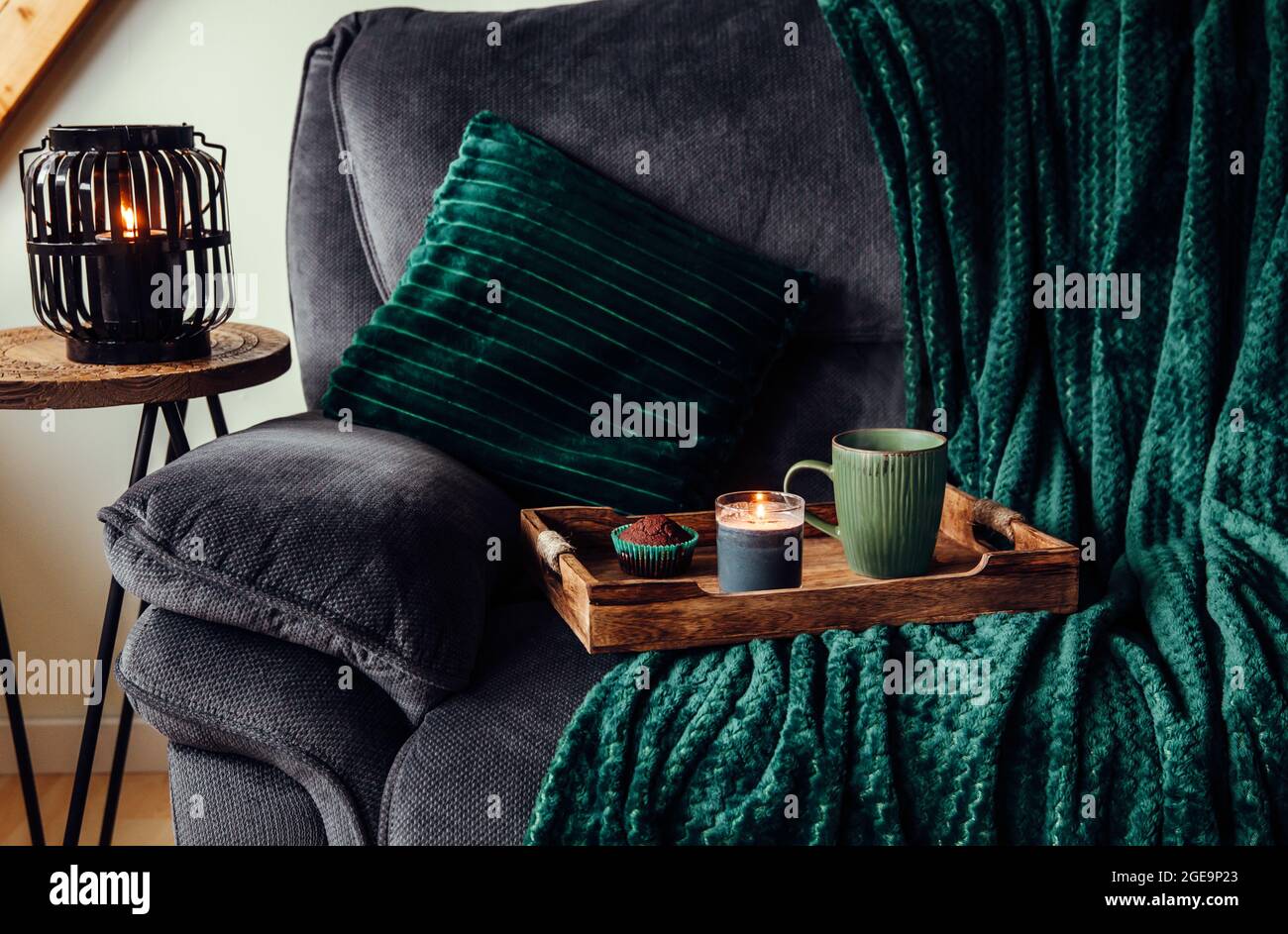 Moderne Herbsthygge im Wohnzimmer. Dunkelgrüne Innenelemente, weiche Kissen, karierten Sofa mit Schokoladenmuffin, Aroma-Trinkbecher auf Holztablett Stockfoto