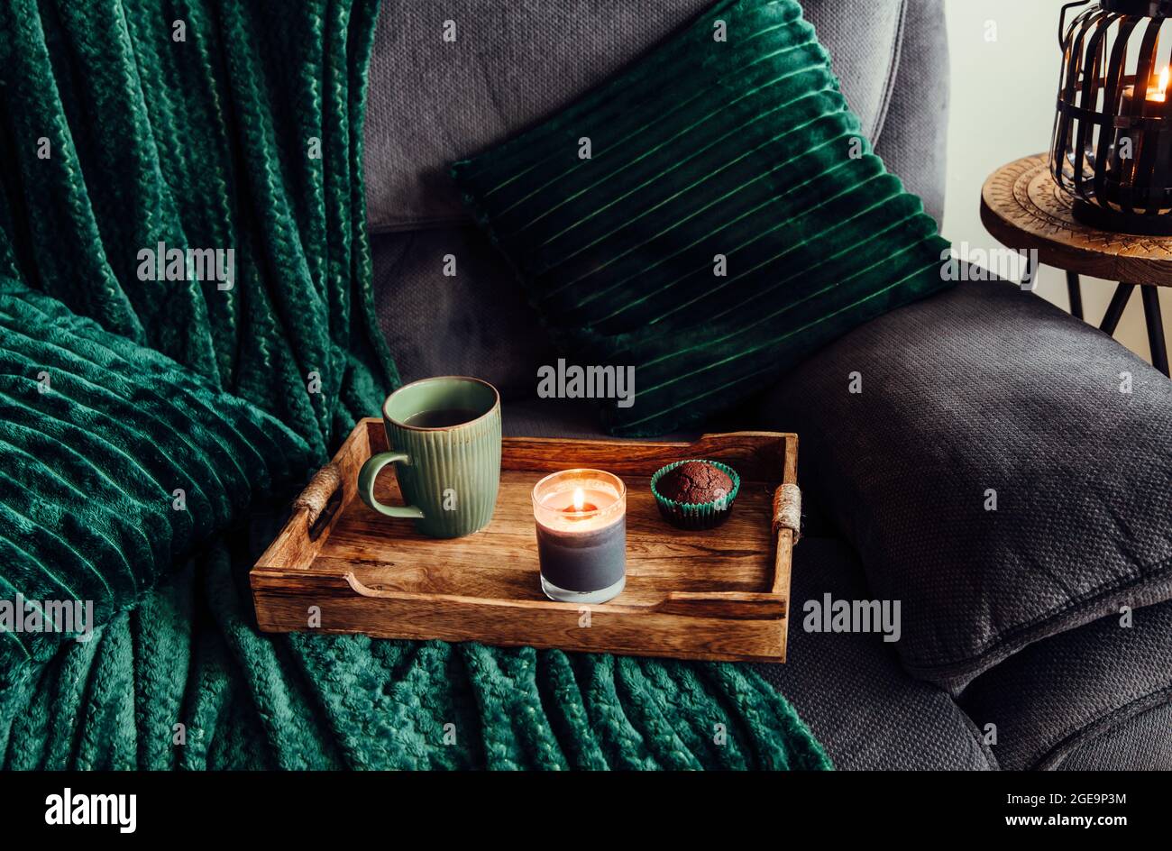 Moderne Herbsthygge im Wohnzimmer. Dunkelgrüne Innenelemente, weiche Kissen, karierten Sofa mit Schokoladenmuffin, Aroma-Trinkbecher auf Holztablett Stockfoto