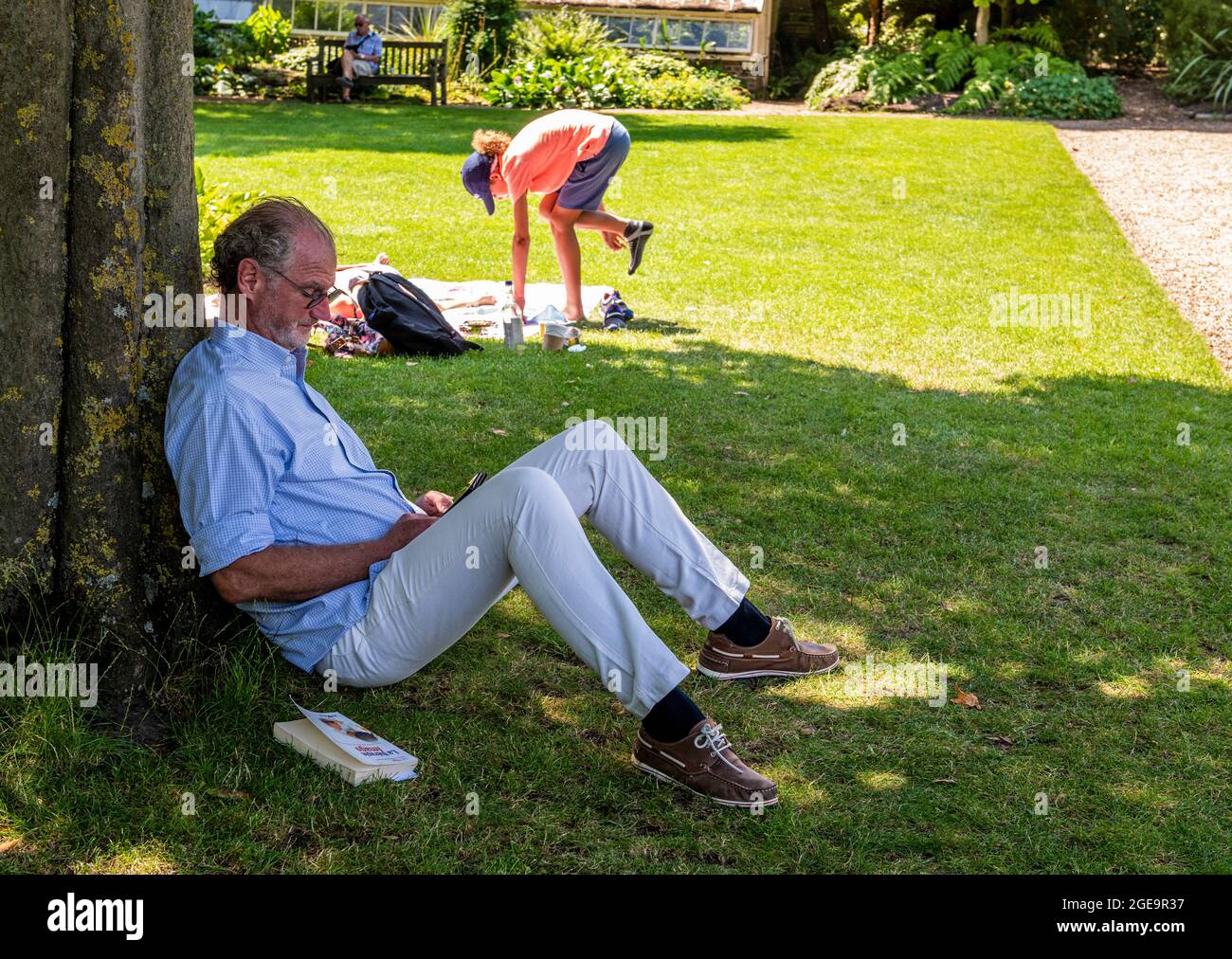Ein Mann, der im Chelsea Physic Garden liest und sich an einem großen Baum ausruht, während eine Frau im Hintergrund versucht, sich auszubalancieren, um ihre Schuhe anzuziehen. Stockfoto