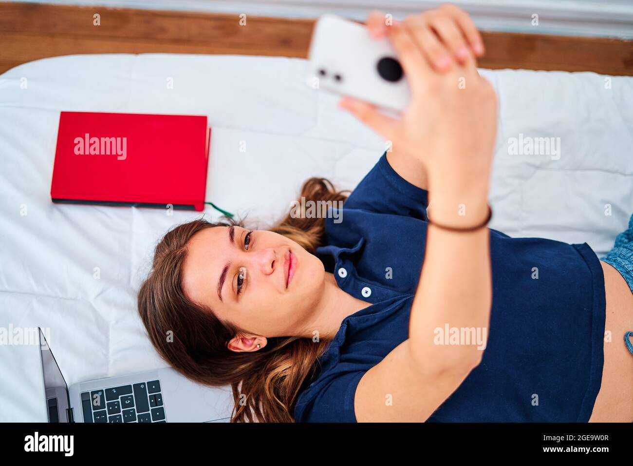 Von oben eine positive junge Studentin, die auf dem Bett liegt und Selfie auf dem Smartphone nimmt, während sie während eines Remote-Online-Studiums zu Hause Pause hat Stockfoto