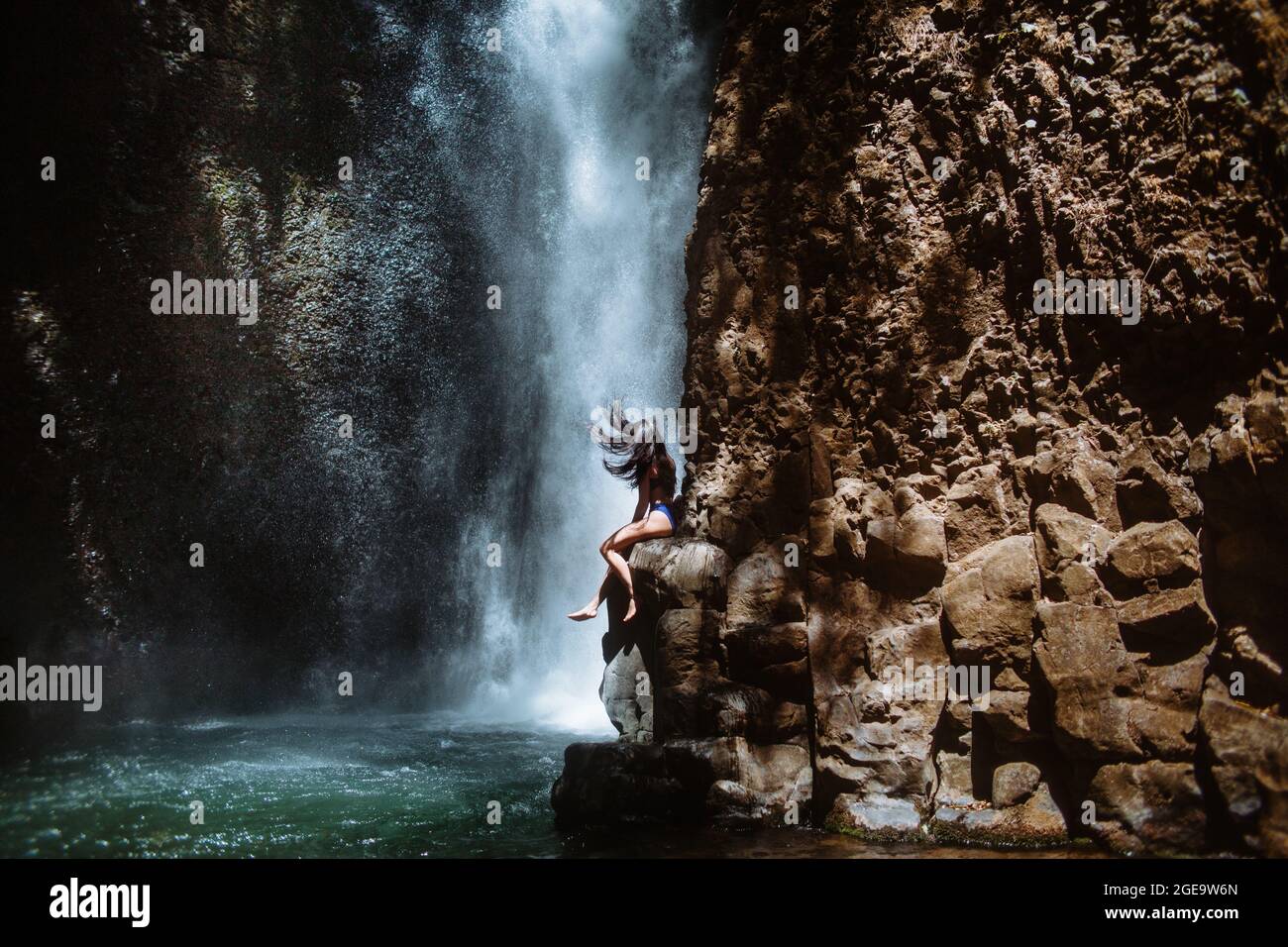 Seitenansicht der unkenntlichen weiblichen Reisenden, die sich entspannen und die frische Luft genießen, während sie am sonnigen Tag i auf einem rauen felsigen Hang in der Nähe eines plätschernden Wasserfalls sitzen Stockfoto