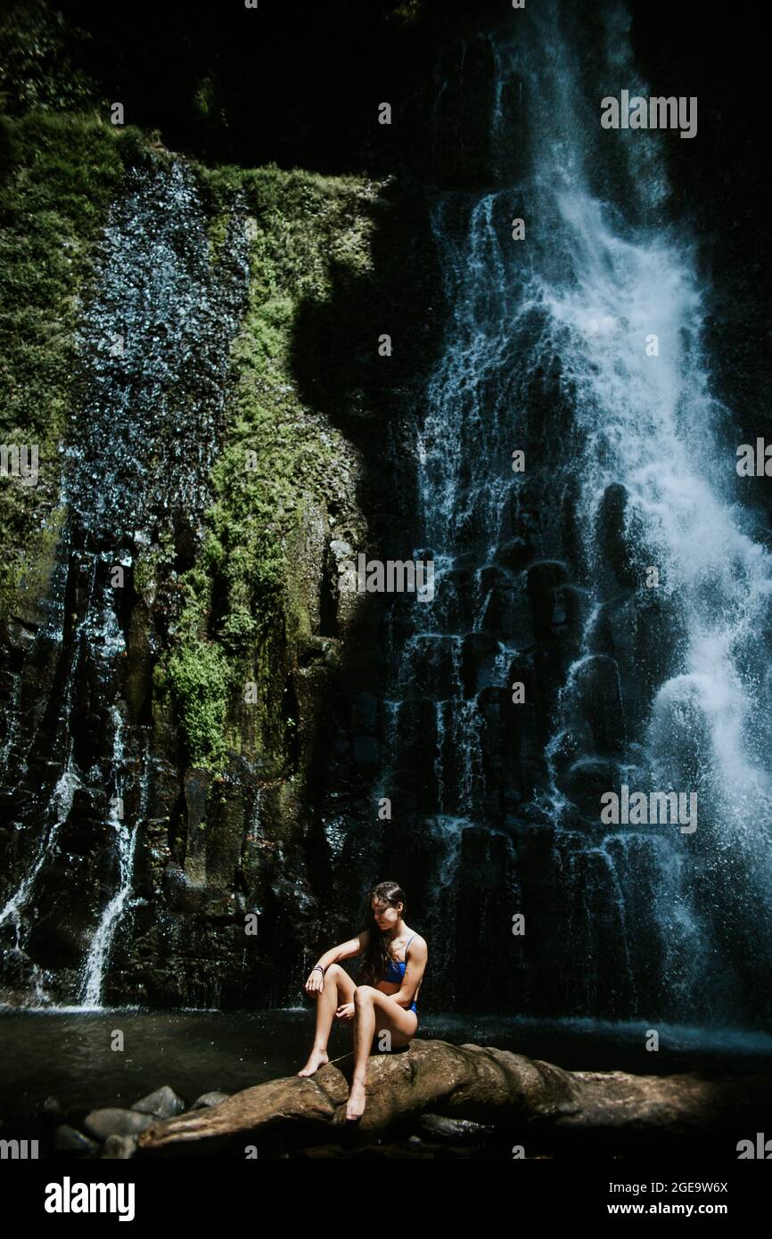 Junge barfuß-Abenteurerin im Bikini, die an sonnigen Tagen in Los Chorros Mun auf Stein gegen steilen Felsen mit grünem Moos und plätscherndem Wasserfall sitzt Stockfoto