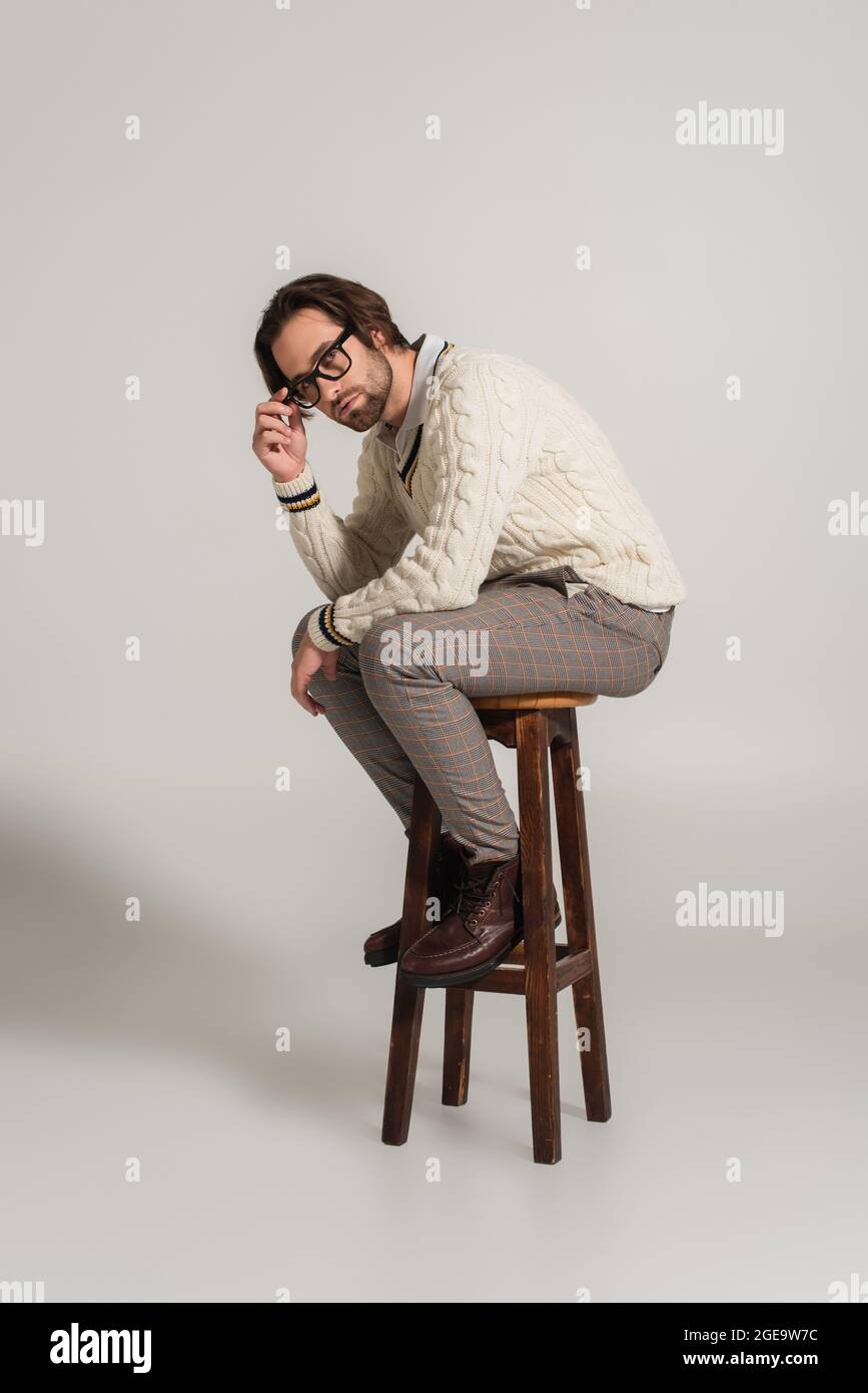 Mann in weißem Pullover und Hose, der die Brille anpasst, während er auf grauem Hintergrund sitzt Stockfoto