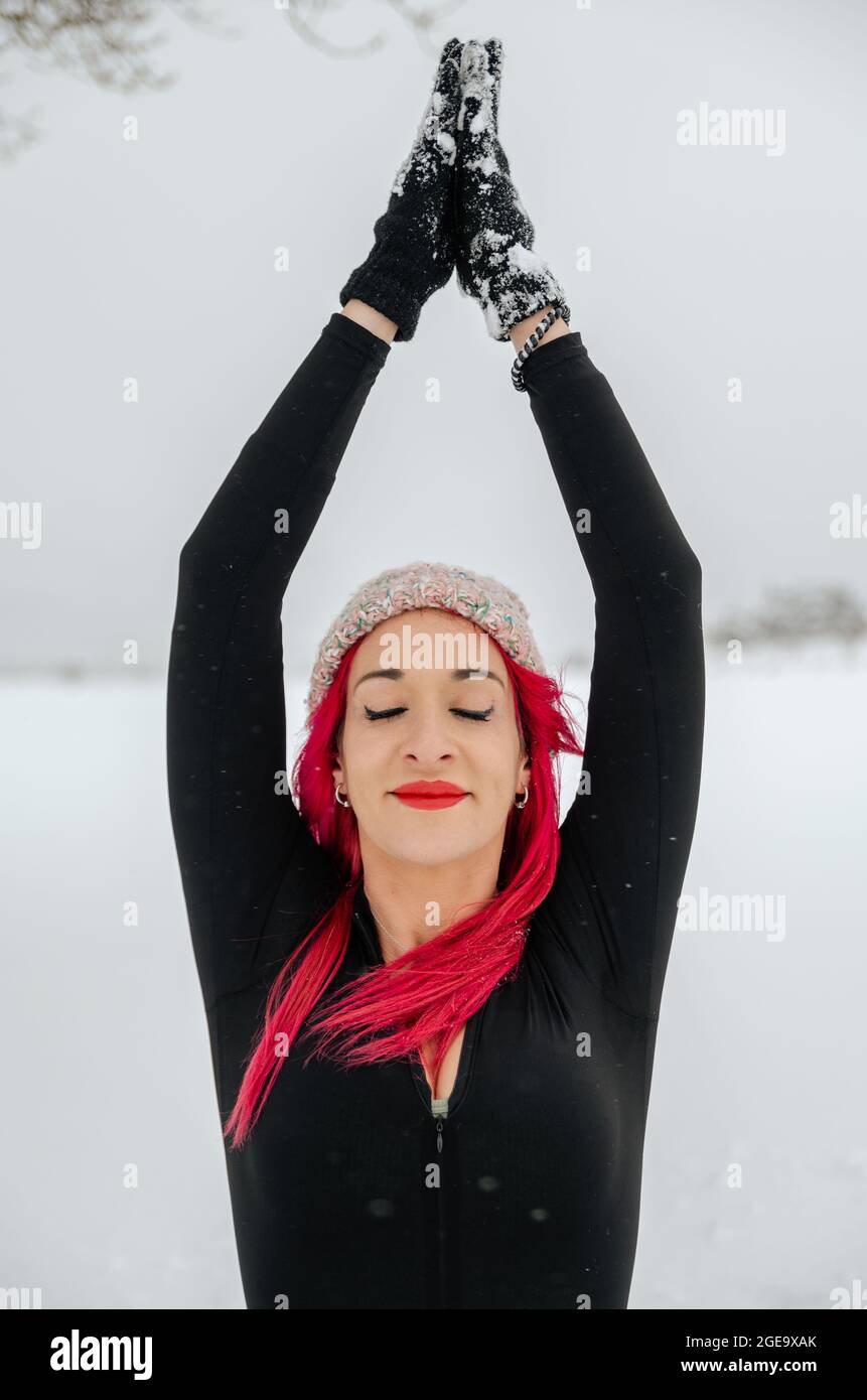 Inhalt Frau mit rosafarbenem Haar praktiziert Yoga in Gomukhasana, während sie mit Namaste-Geste im schneebedeckten Winterfeld sitzt Stockfoto