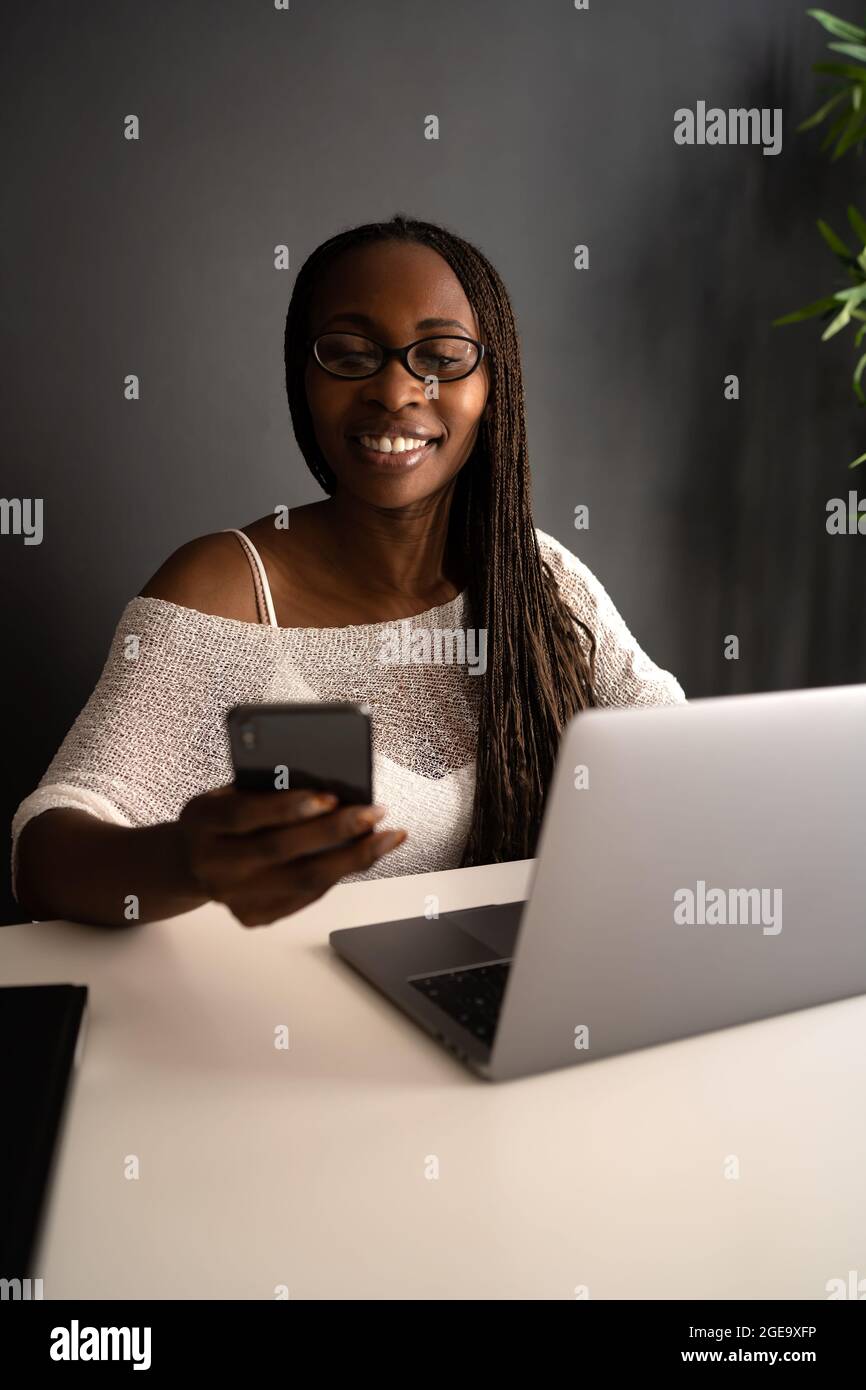 Fröhliche afroamerikanische Freiberuflerin, die am modernen Arbeitsplatz am Tisch sitzt und am Mobiltelefon surft, während sie an einem Projekt außerhalb von zu Hause arbeitet Stockfoto