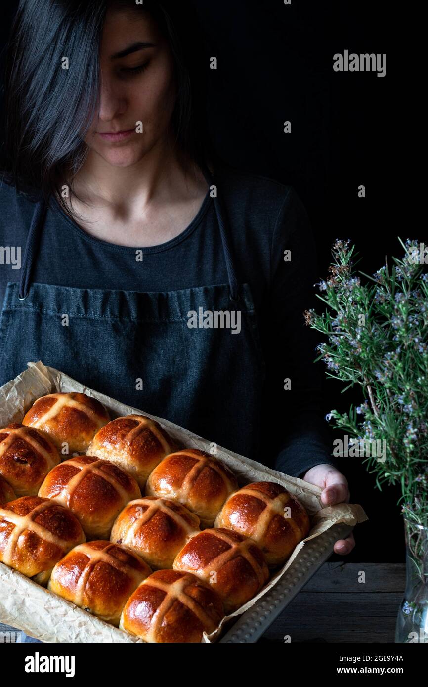 Weibliche Bäckerin hält Blätterteig frisch gebackene, heiße Kreuzbrötchen auf dem Backblech Stockfoto