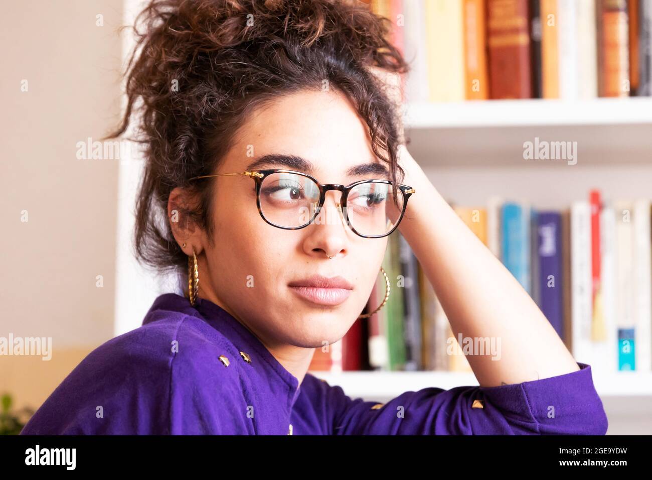 Junge hispanische Hündin mit lockigem Haar in Freizeitkleidung und Brille mit Kreolen, die den Kopf an der Hand lehnen und die Kamera betrachten, während sie in der Nähe von Boo stehen Stockfoto