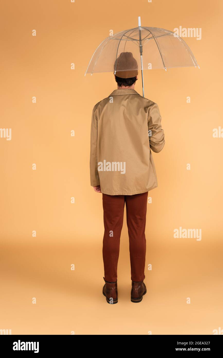 Rückansicht des Mannes in brauner Hose und Jacke, der unter transparentem Schirm auf beige steht Stockfoto