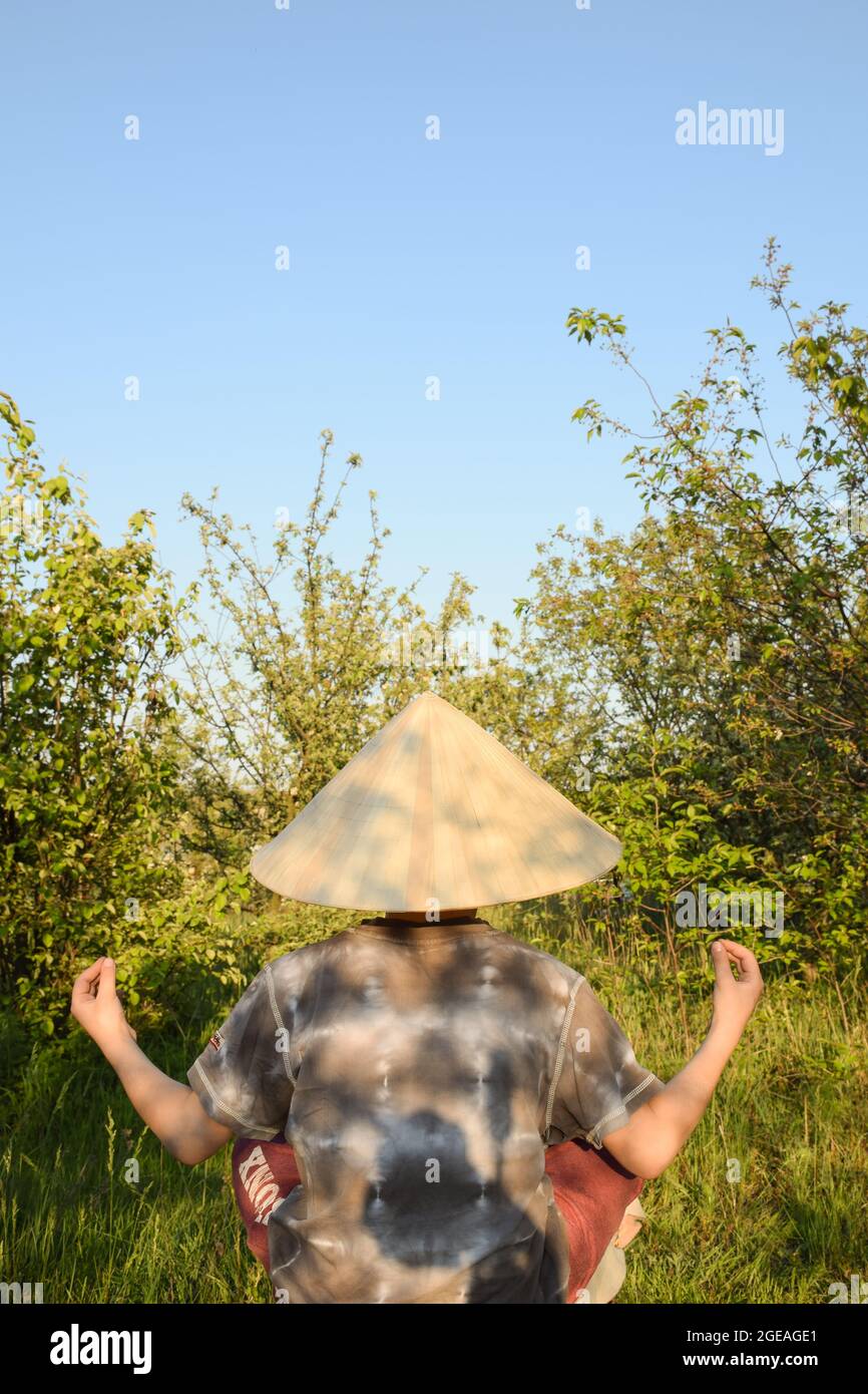 Der kleine Junge sitzt mit seinem Rücken in einer Zen-Pose in einem chinesischen Kopfschmuck auf einem Hintergrund aus Bäumen und blauem Himmel Stockfoto