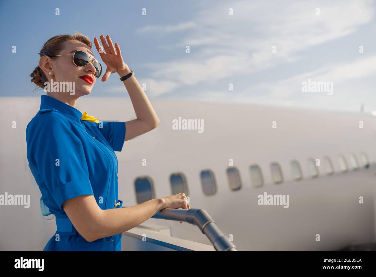 Porträt einer eleganten Luftverkehrsleiterin in blauer Uniform und einer Sonnenbrille, die die Hand am Kopf hält und weit weg schaut und tagsüber auf der Lufttreppe steht Stockfoto