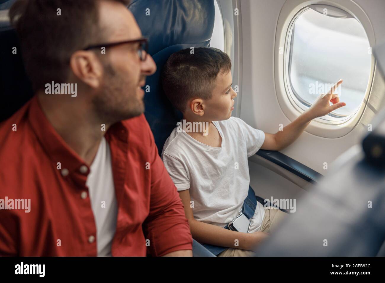 Neugieriger kleiner Junge, der aus dem Fenster schaut, während er mit seinem Vater im Flugzeug unterwegs ist Stockfoto