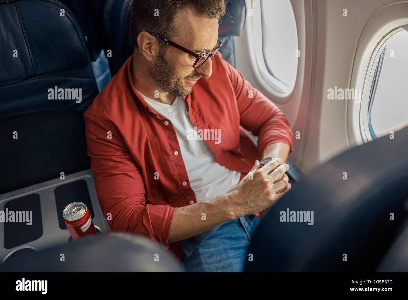 Ein hübscher kaukasischer Mann in legerer Kleidung und Brille, der die Ankunftszeit auf seiner Uhr überprüft und auf dem Flugzeug in der Nähe des Fensters sitzt Stockfoto