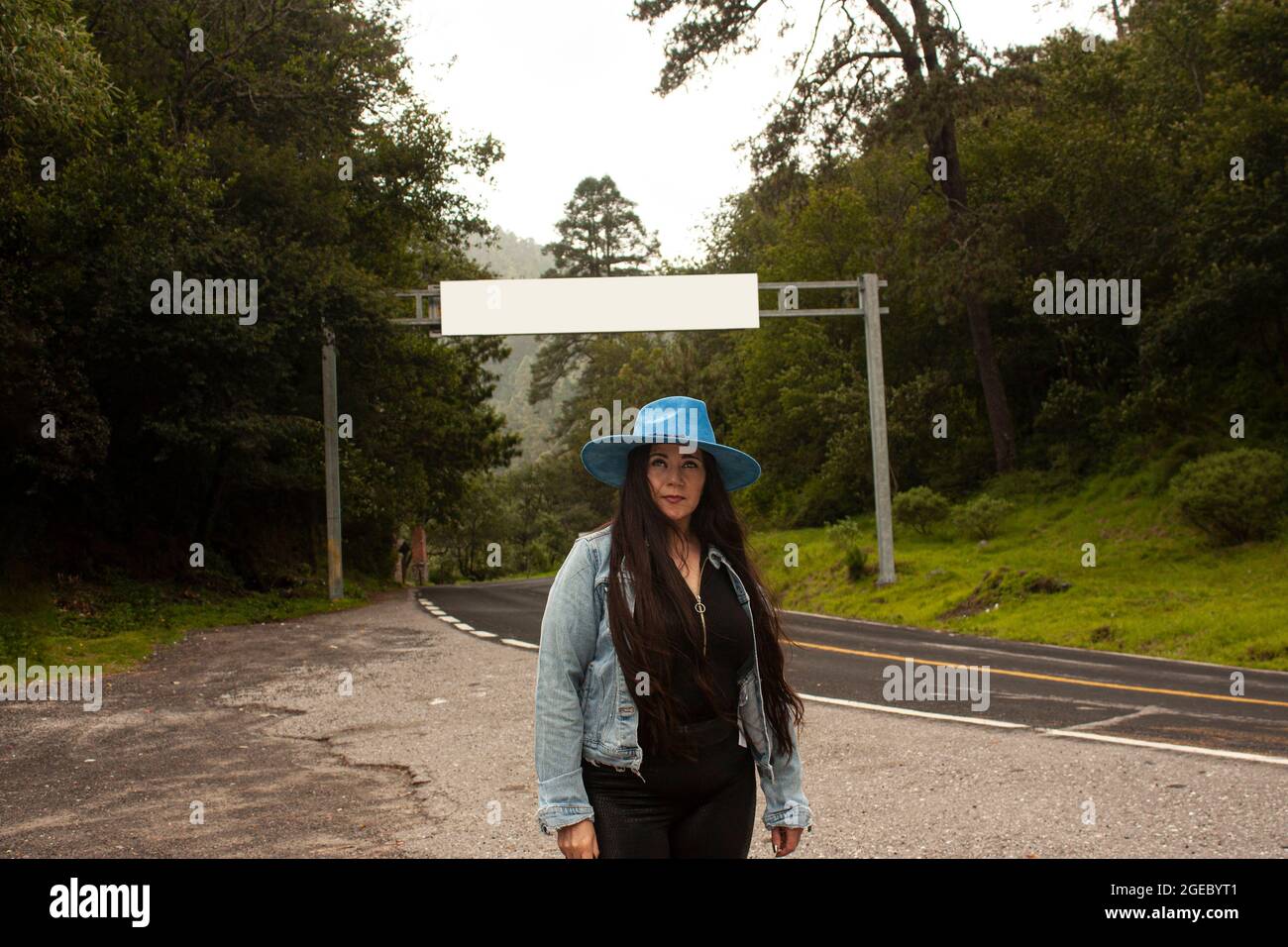 Frau, die auf der Straße ohne Richtung, umgeben von Wald, trägt Denim-Jacke und Hut in Mexiko Stockfoto