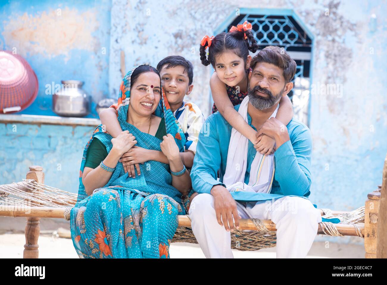Porträt einer glücklichen ländlichen indischen Familie, die auf die Kamera schaut, während sie im Dorfhaus auf einem traditionellen Bett sitzt, umarmen kleine Tochter und Sohn Vater und mothe Stockfoto