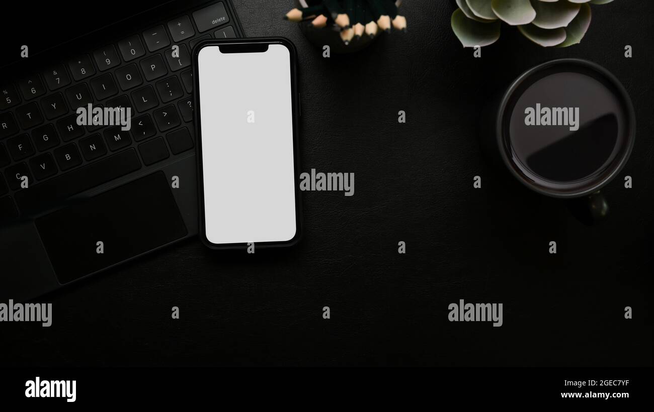 Moderner Arbeitsplatz, Smartphone-White-Screen-Mockup auf schwarzer magischer Tastatur, Bleistifte, Pflanze, Kaffeebecher, schwarze Oberfläche, Draufsicht, oben Stockfoto