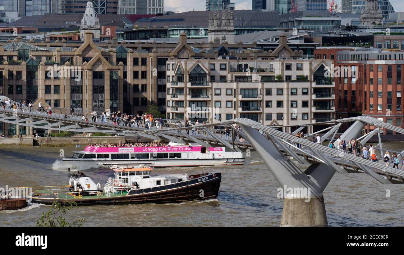 London, Greater London, England, August 10 2021: Boote fahren unter der Millennium Bridge an der Themse mit Gebäuden im Hintergrund. Stockfoto