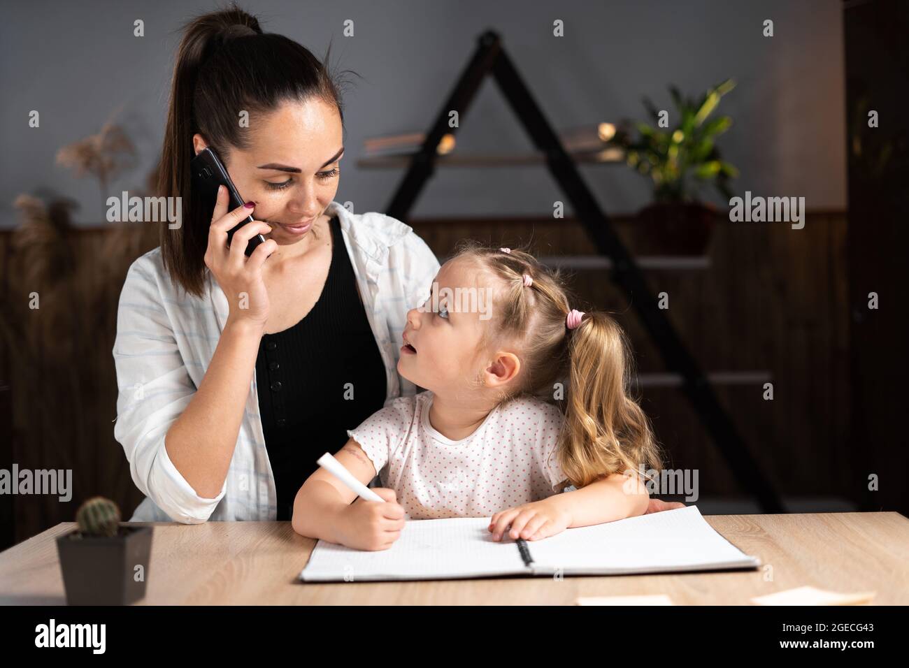 Eine junge kaukasische Mutter und ihre Tochter sitzen abends zu Hause am Tisch und machen gemeinsam Hausaufgaben. Ein junges Kindermädchen hilft dem Kind d Stockfoto