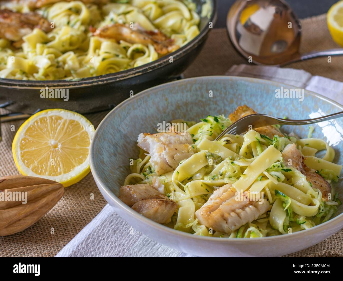 Fischgericht mit Pasta, Zuccini und weißer Sauce serviert auf einem Teller mit Gabel auf Küchentisch Hintergrund mit Pfanne und Schöpfkelle Stockfoto