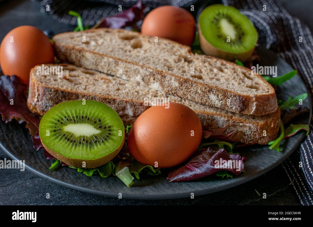 Ein Teller mit vegetarischen Speisen wie Bio-gekochten Eiern, Sauerteig und frischen halbierten Kiwis. Nahaufnahme auf dunklem Hintergrund Stockfoto