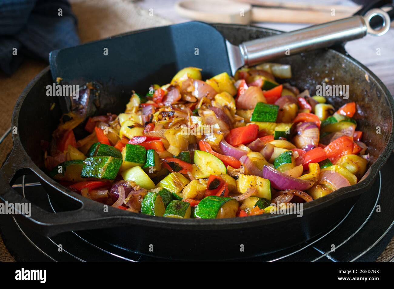Gebratenes Gemüse in einer rustikalen gusseisernen Pfanne serviert. Mit roten Zwiebeln, Zucchini, roten Paprika und Knoblauch. Serviert auf einem Holztisch Stockfoto