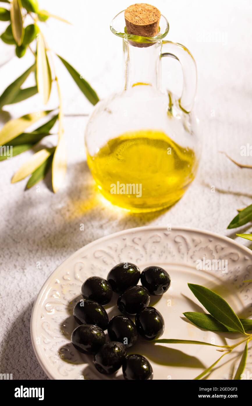 Ein Glas mit nativem Olivenöl, strukturierte Platte mit schwarzen Oliven und einem Zweig von Olivenbaum Stockfoto