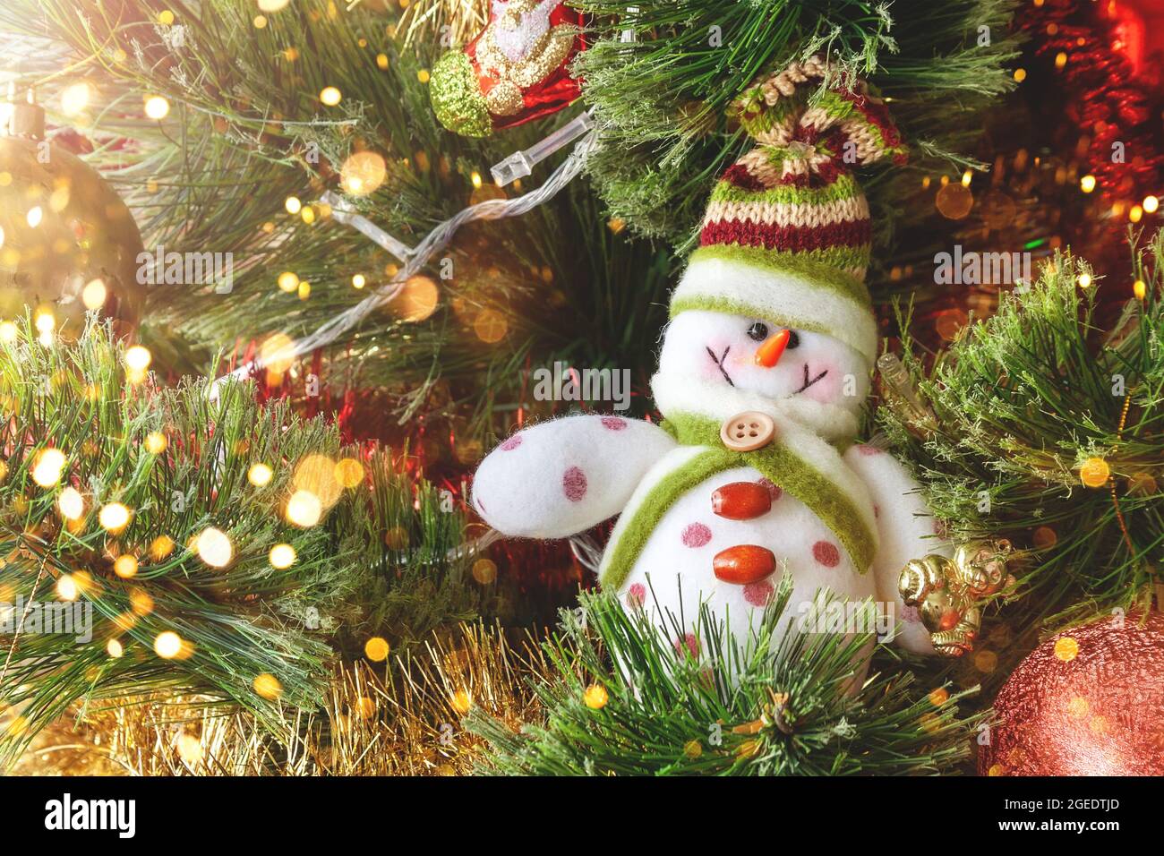 Lächelnder Schneemann aus Textil auf dem Weihnachtsbaum mit bunten, unscharfen Lichtern. Selektiver Fokus. Weihnachtskarte mit Dekorationen für den Silvesterabend Stockfoto