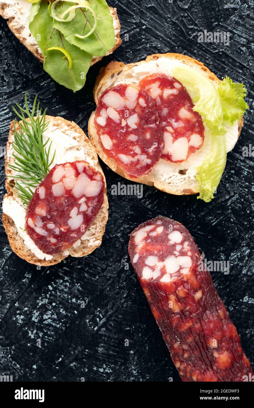 Wurst und drei Sandwiches oder Snacks mit ihnen auf einem schwarzen strukturierten Hintergrund. Weicher Fokus. Flach liegend Stockfoto