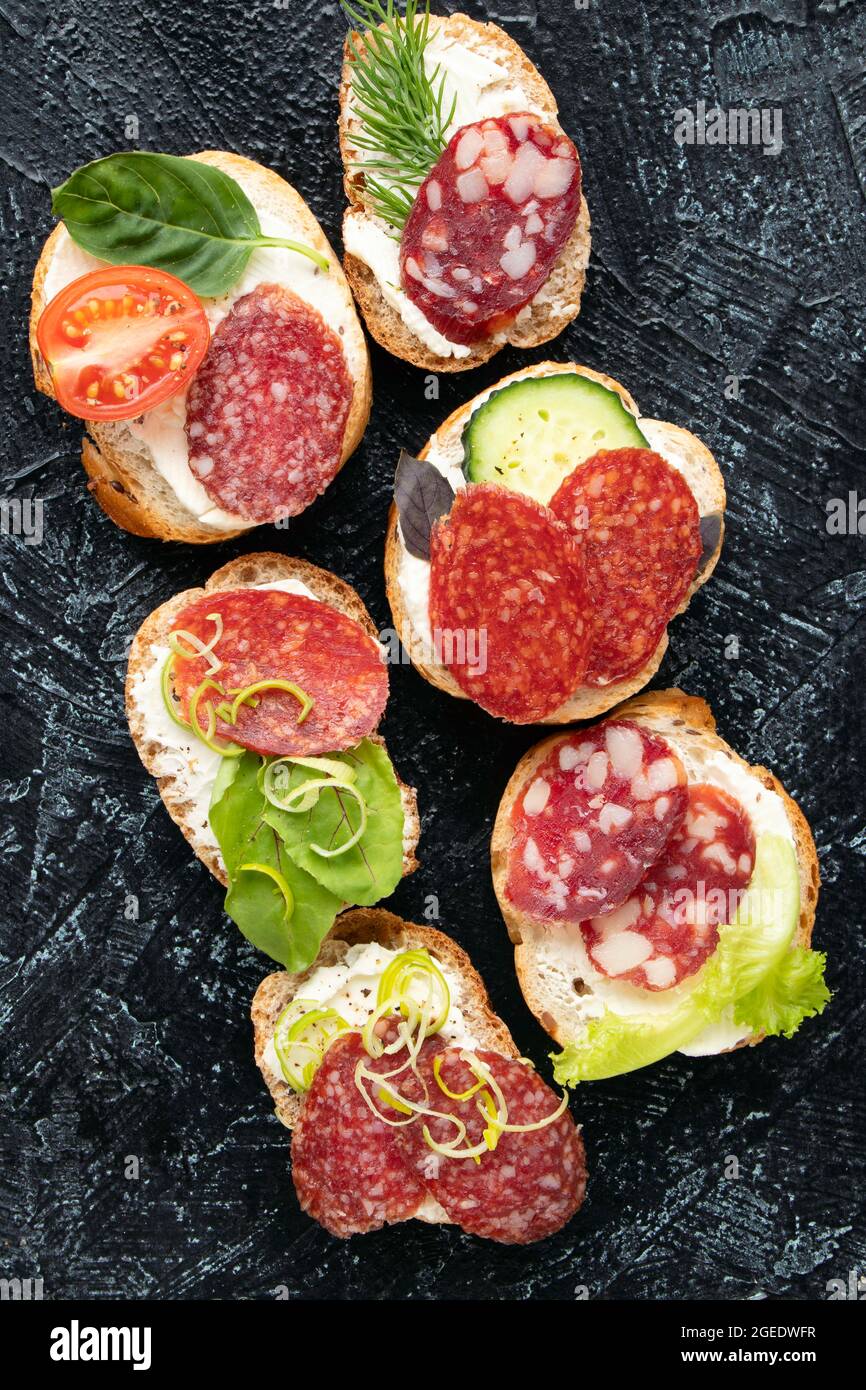 Arten Sandwiches oder Snacks mit verschiedenen Sorten Wurst und Gemüse auf schwarzem Hintergrund. Weicher Fokus. Draufsicht. Vertikale flache Lage Stockfoto